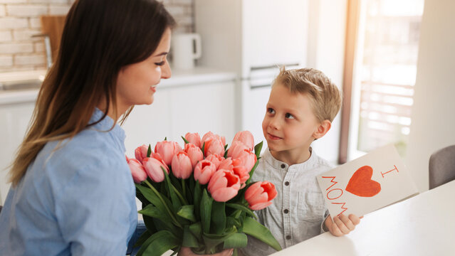 Young woman with her little son and greeting card for Mother's Day at home. Happy woman receiving flowers and greeting card from her son at home. Mother's day celebration