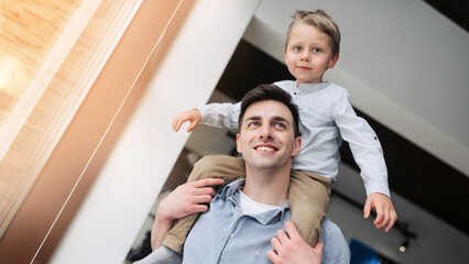 A father with a young son are fooling around while sitting on the sofa. The boy sits on his father's shoulders. They're laughing