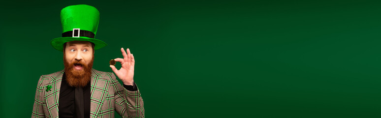 Excited bearded man in hat holding coin during saint patrick day isolated on green, banner.