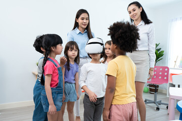 A group of multi-ethnic preschool children, boys and girls, standing around a little boy in a virtual reality simulator headset, playing in a classroom under the supervision of two beautiful teachers.