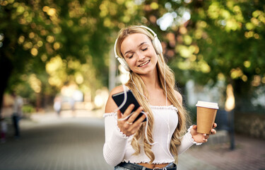 Cheerful teenager student girl walking on the streets with a takeaway paper coffee cup in her hand and listening to music with headphones from her smart phone