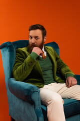 Trendy bearded model looking at camera while sitting on armchair on red background.