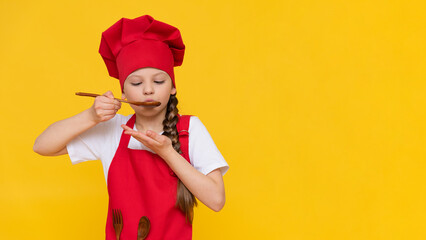 A child chef tastes a delicious dish with a spoon in a red apron and a hat looks at your advertisement on a yellow isolated background.