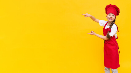 The chef child points sideways at your advertisement in a red apron and cap on a yellow isolated background. Copy space. A place for advertising.