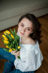 Spring portrait of young beautiful woman with blue eyes and curly hair with bouquet of yellow tulips in home interior. Happy model in jeans and white shirt. Mothers Day.