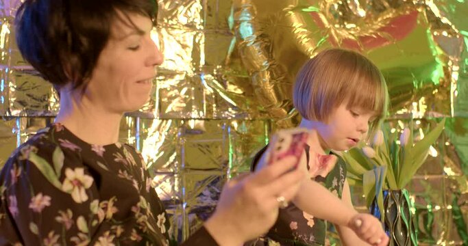 Attractive mom takes a selfie photo with her daughter on her birthday. She holds the phone, bows her head to the child. Golden background.
