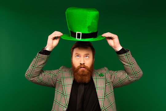 Bearded man in jacket looking at hat during saint patrick celebration isolated on green.