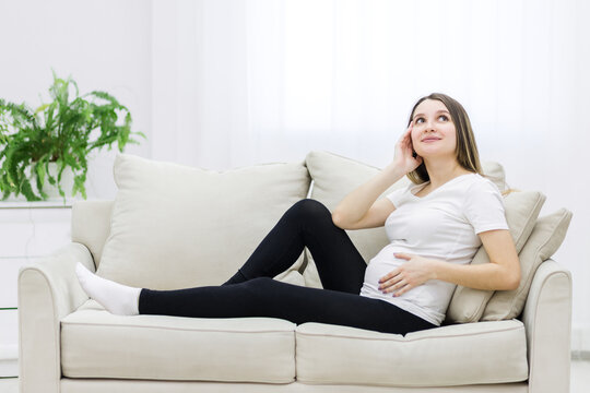 Young pregnant woman thinking about something on white sofa.