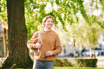 Outdoor sunny portrait of handsome happy young red-haired man in spring park, wearing brown fuzzy fleece sweater and backpack - 572749321
