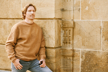 Naklejka na ściany i meble Outdoor fashion portrait of handsome red-haired man posing next to brown wall, wearing beige fuzzy fleece sweater