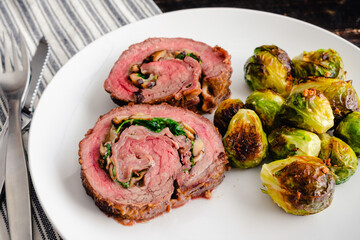 Stuffed Flank Steak with Prosciutto and Mushrooms Dinner: Slices of steak roulade served on a plate with roasted Brussels sprouts