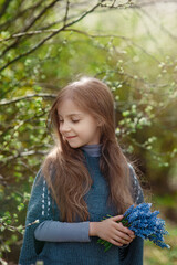 Fototapeta na wymiar Pretty little happy smiling girl with long hair and bouquet of blue flowers in spring park with greenery and sunshine in background, cosy warm child outdoor portrait, tenderness and beauty of nature