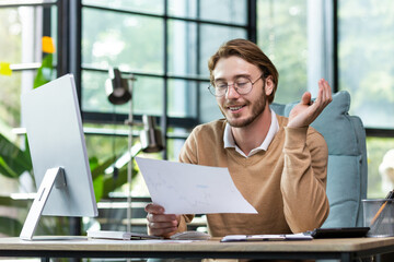 A young male student sits in the office and holds documents in his hands, studies, works at the computer, smiles.