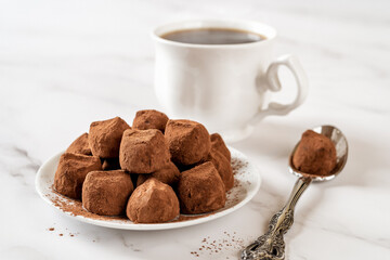 Chocolate truffle balls on a white plate. Heap of dark chocolate candies in cocoa powder, cup of coffee and spoon on a marble table. Sweet food and gourmet confection concept. Macro.