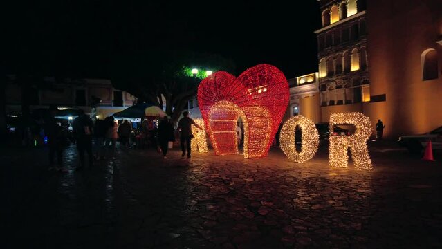 Pareja paseando frente a letras en 3d  en forma de Corazon, las letras estan iluminadas por la noche, y dicen la palabra amor 
