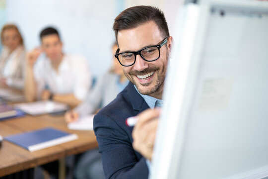 Close up portrait of man in formal-wear and eyeglasses using pen and writing graphs and data on flip chart in front of his business partners at conference or meeting room.