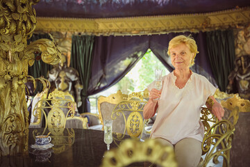 Mature attractive woman traveler sitting alone on the terrace of coffee shop in beautiful park in Bulgaria and drinking champagne. Active life of the elderly in retirement, active seniors