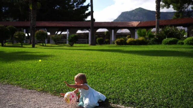 Little girl with a ball in her hands walks on a green lawn and falls