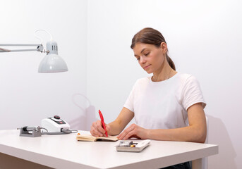 Beautiful nail beautician makes notes in notebook with pen, sitting at workstation, table in nail beauty treatment salon. Professional tools are on white table. Manicurist's routine. Horizontal plane