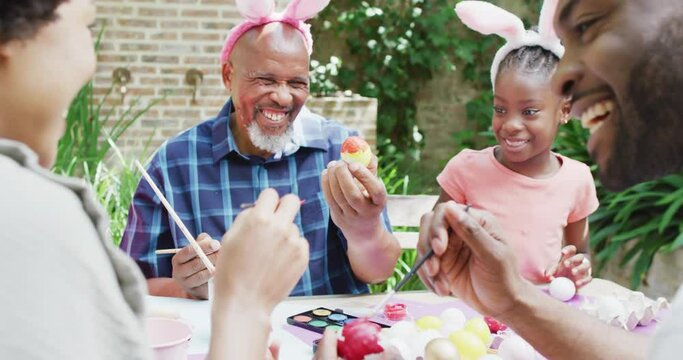 Happy african american parents, daughter and grandfather painting easter eggs in garden, slow motion