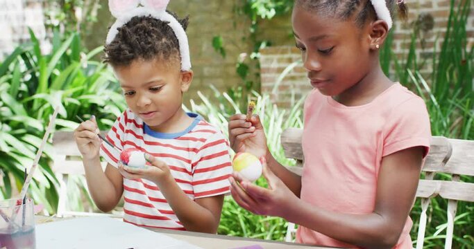 Happy african american brother and sister in bunny ears painting eggs for easter, slow motion