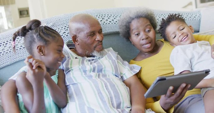 Happy african american grandparents, grandson and granddaughter on couch using tablet, slow motion