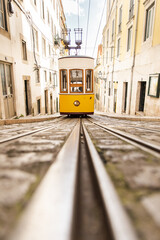 Vertical shot, an old vintage yellow electric tram in the style of the Lisbon funiculars rides on rails on a narrow cobblestone street between low-rise simple European houses on a sunny day - obrazy, fototapety, plakaty