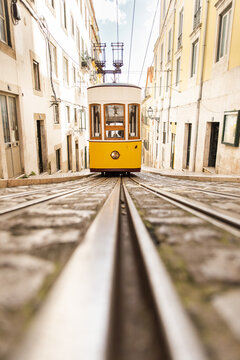 Vertical shot, an old vintage yellow electric tram in the style of the Lisbon funiculars rides on rails on a narrow cobblestone street between low-rise simple European houses on a sunny day