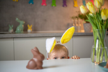 A cute little boy wearing bunny ears on Easter day wants to eat a chocolate easter bunny. A child plays egg hunt for Easter. Charming child celebrates Easter at home.