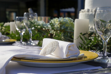 Elegant table setting decorated with green leaves boho-inspired centerpiece, shells and white linen cloth for a engagement party, wedding reception or other outdoor summer event.