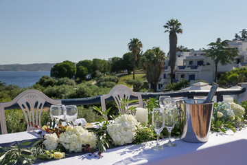 Elegant table setting decorated with white flowers and green leaves centerpiece and white linen cloth for a engagement party, wedding reception or other outdoor summer event with sea view