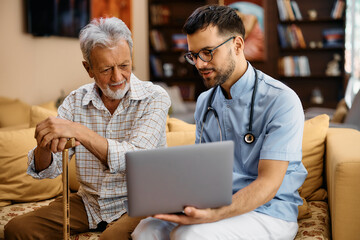 Obraz premium Senior man using laptop with help of male caregiver during home visit.