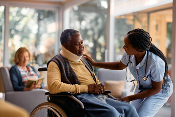 Caring African American nurse talks to senior man in wheelchair at nursing home.