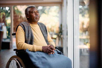 Thoughtful black senior man in wheelchair looks through window at residential care home.