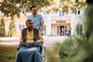 Young healthcare worker pushes senior black man in wheelchair through nursing home's park.