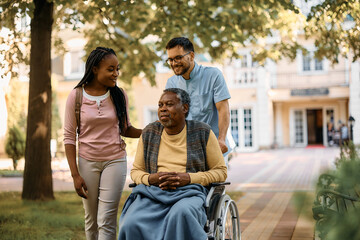 Smiling black senior in wheelchair enjoys with his daughter and caregiver in park at nursing home.