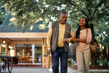Happy black woman walks with her senior father while visiting him at nursing home.