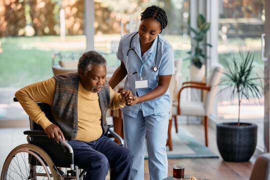 Young black nurse assists senor man to get up from wheelchair at nursing home.