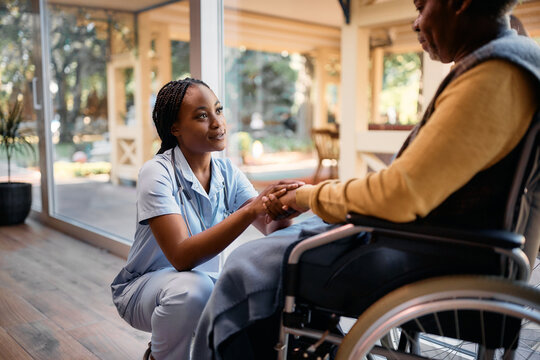 Caring black nurse comforting senior man in wheelchair at nursing home.