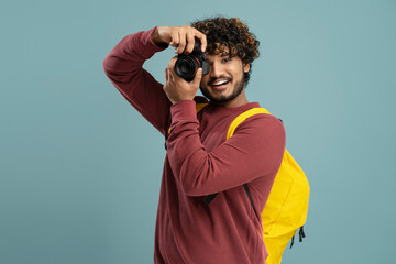 Smiling young Indian man with backpack, taking photo on his modern digital camera on blue background
