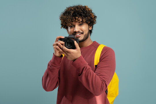 Happy curly haired Asian guy photographer, journalist or traveler with yellow backpack, holding digital camera and smiling looking at camera, before taking photos isolated on blue background