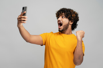 Overjoyed young curly bearded Indian man in yellow t-shirt holding a modern mobile phone in winning lottery, reading good news, isolated over gray background. Guy sports betting celebration success 