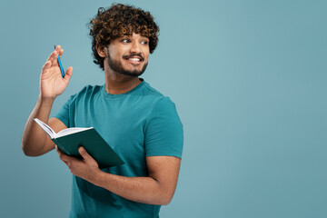 Smiling handsome young Indian man with notepad looking aside isolated over blue color background