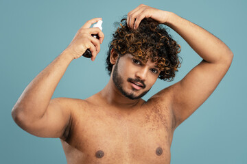Smiling handsome Hindu man with naked torso, applying hair care spray isolated over blue background