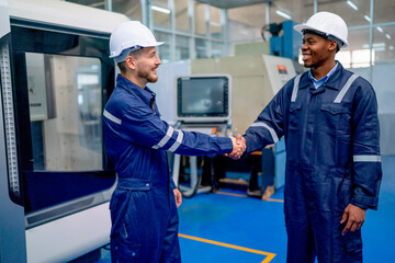 Caucasian worker and African American technician shake hand with smiling face in front of factory robotic machine during work in workplace.