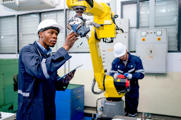 African American engineer or technician worker hold part of robotic arm and check  the function of machine and co-worker support in the back in factory workplace.