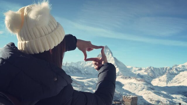 Back view of young traveler girl standing over snowy mountain peaks and making frame gesture by hands with Matterhorn Switzerland, Winter traveling scene and planning.