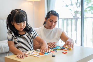 Fototapeta premium Medium shot portrait of Asian girl learning English alphabet by playing jigsaw puzzle in living room at home, near her mom. Child care, family, mother and daughter concept