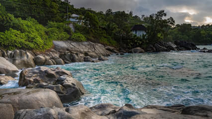 The waves of the turquoise ocean beat against the coastal granite boulders. Foam on the water. Houses are visible through the lush vegetation. The clouds in the sky are illuminated by the sun. 