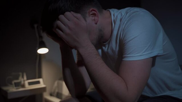 Bad thoughts and dreams. Upset young caucasian man presses his hands to his head and cannot sleep in bed at night. Side effects of coronavirus insomnia, depression and melancholy. Sleep disorder.